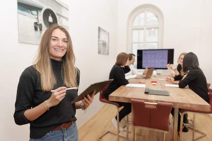 Eva steht im Vordergrund mit einem Tablet in der Hand, mehrere Mitarbeiter sitzen im Hintergrund an unserem Meetingtisch.