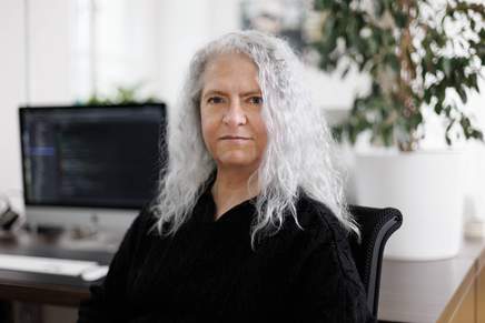 Mitarbeiterin Portrait: Frau mit langen, weißen Haaren. Computerbildschirm steht im Hintergrund.