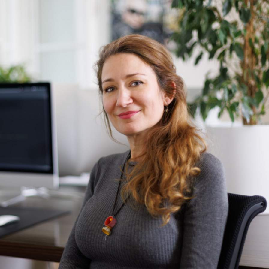 Mitarbeiterportrai von Cristina. Dame mit langen braunen Haaren. Im Hintergrund sieht man unser Büro mit einem PC-Bildschirm und einer großen Pflanze.