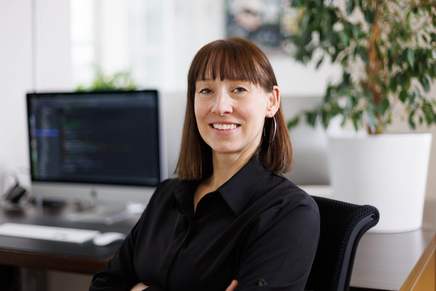 Mitarbeiterin Portrait: Frau mit schulterlangen, dunklen Haaren. Computerbildschirm steht im Hintergrund.